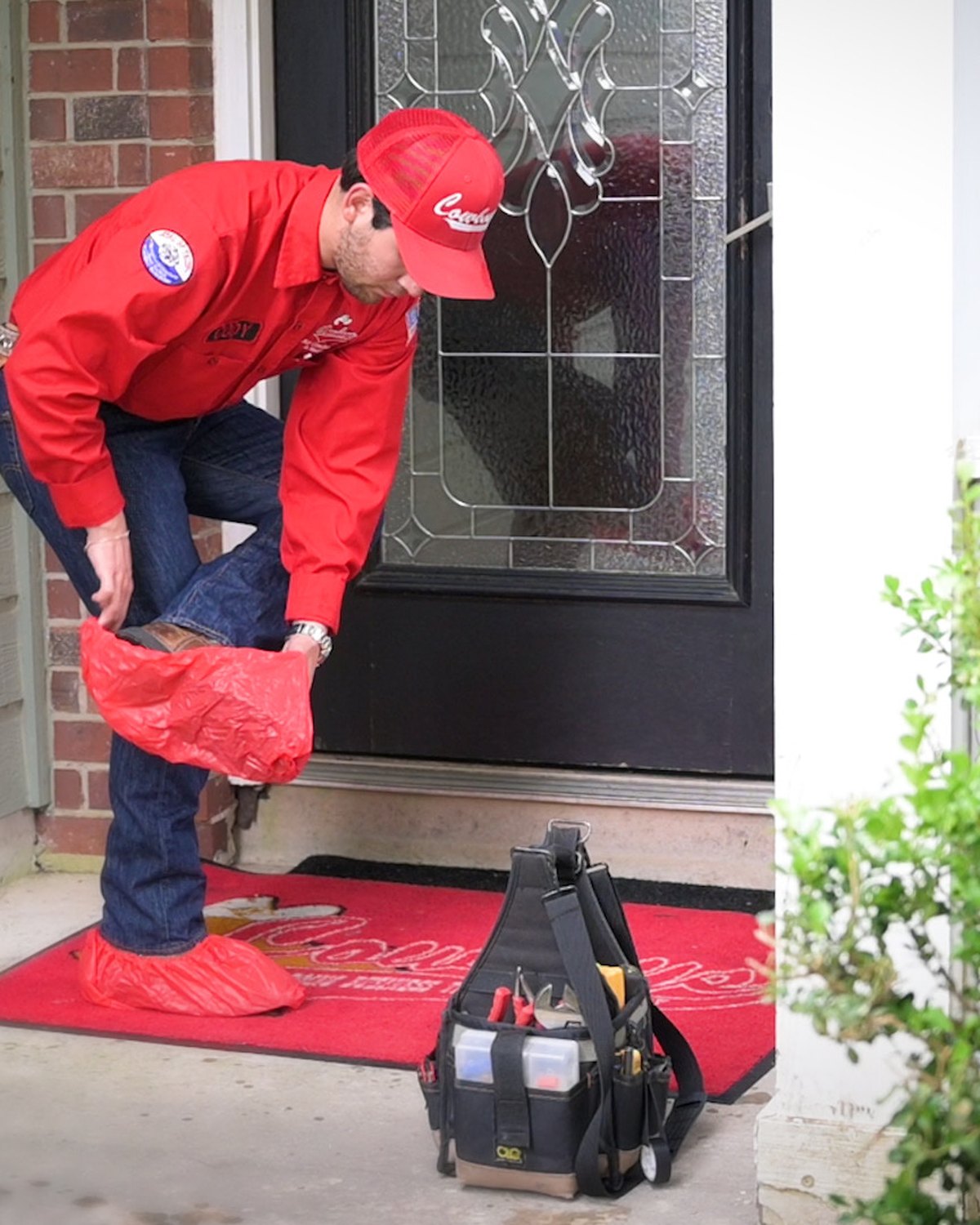 A Cowboy's tech pulling on red boot covers at a customer's front door, tool bag down